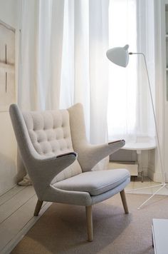 a white chair sitting in front of a window next to a book shelf and lamp