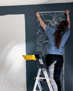 a woman is painting the wall with blue paint and she is standing on a ladder