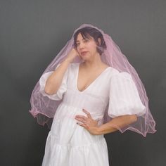 a woman in a white dress is wearing a pink veil and holding her hand to her face