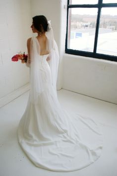 a woman in a white wedding dress standing by a window with her back to the camera