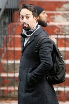 a man with a beard wearing a coat and scarf walks down the street in front of some stairs