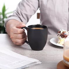 a man holding a black coffee cup in his hand while sitting at a table with food