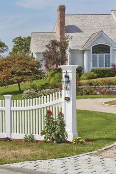 a white picket fence in front of a house with flowers and shrubs around the gate