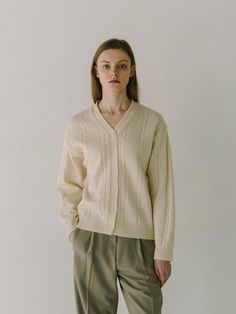 The person in the photograph is wearing a cardigan with a deep V-neckline, which gives it a relaxed and comfortable feel. The cardigan showcases cable knit details along the front, adding a classic and textural element to the design. It features a button-down front with small, subtle buttons that blend into the overall color scheme. The ribbed cuffs and hem offer a snug fit, and the cardigan seems to be made from a material that provides warmth and softness.- The cable knit design creates a timeless aesthetic, indicating that this garment could be a staple in a classic wardrobe.- The V-neckline is versatile, allowing the garment to be styled over various types of clothing, from collared shirts to simple tees.- The neutral color of the cardigan suggests that it could be easily paired with o V-neck Cable Knit Outerwear For Layering, Classic Cable Knit Cardigan For Fall, Beige Cable Knit V-neck Cardigan, Beige V-neck Cable Knit Cardigan, Elegant Beige Cable Knit Cardigan, Chic Cable Knit V-neck Cardigan, Classic Cable Knit Cardigan For Work, Classic Cable Knit Cardigan For Spring, Classic Spring Cable Knit Cardigan