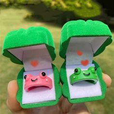 two green frog rings with hearts and eyes are in a small open box on the grass