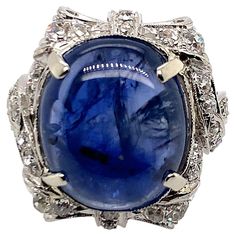 The bold design set with a cabochon sapphire weighing approximately 14.00 carats, framed by elaborate ribbon bows and sash motifs set with round diamonds, in 18k white gold, size 5 1/2 Weight: 9.2 grams Sapphire measuring 15.19 x 12.90 x 5.56 mm, of a medium light tone of slightly violetish blue colour, noticeably included, good cut Diamonds estimated weight 0.75 carat, G-I colour, VS-I1 clarity Good condition Domed Ring, Bold Design, White Gold Ring, Blue Colour, Design Set, Gem Stone, Sapphire Diamond, Ribbon Bows, White Gold Rings