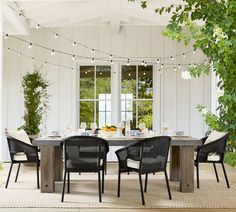 an outdoor dining table with chairs and lights strung from the ceiling, in front of a white house