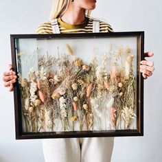 a woman holding up a framed photo with dried flowers in the middle and grass on the bottom