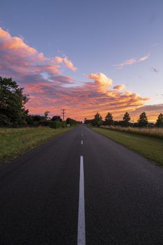 an empty road in the middle of a grassy field with trees on both sides at sunset