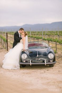 a bride and groom kissing in front of a vintage car on the side of a dirt road