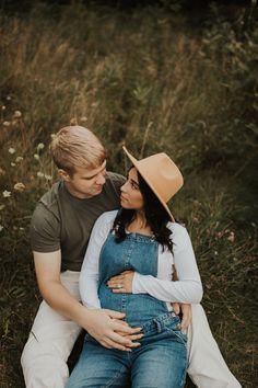 a man and woman sitting on the ground in front of some tall grass with their arms around each other