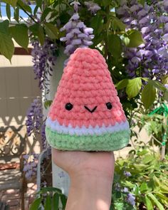 a hand holding up a small crocheted watermelon toy in front of purple flowers