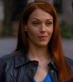 a woman with long red hair wearing a black leather jacket and blue shirt looking at the camera