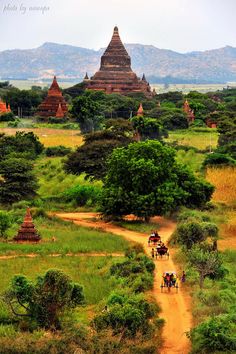 several people riding horses down a dirt road in front of some pagodas and trees