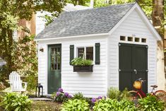 a small white shed with green shutters and window boxes on the side, surrounded by greenery