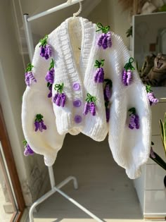 a white sweater with purple flowers on it hanging from a clothes rack next to a potted plant