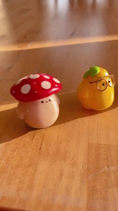 two small toy mushrooms sitting on top of a wooden table next to an orange and a yellow apple