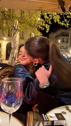 two women hugging each other at an outdoor restaurant with wine glasses on the table in front of them