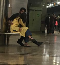 a man and woman sitting on a bench in the middle of a subway station with people standing around