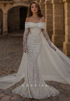 a woman in a white wedding dress standing on a cobblestone walkway wearing an off the shoulder veil