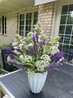 a white vase filled with purple and white flowers on top of a wooden table next to a brick building