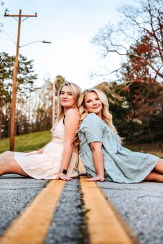 two beautiful young women sitting on the side of an empty road posing for a photo