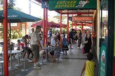 a group of people walking down a sidewalk next to tables with umbrellas over them