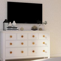 a white dresser with gold knobs in front of a flat screen tv mounted on the wall