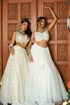 two beautiful women standing next to each other in front of a wooden door wearing white dresses