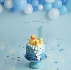 a blue cake with two rubber ducks on top and balloons in the background for a first birthday party