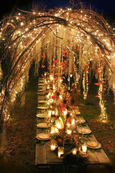 an outdoor dinner table set up with candles and lights on the branches, surrounded by greenery