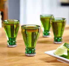 four green glasses sitting on top of a wooden table next to a plate with lime wedges