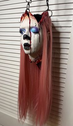 a creepy mask hanging from a hook on a wall with long pink hair and blue eyes