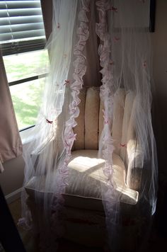 a chair covered in white netting with pink flowers on the back and sides, sitting next to a window