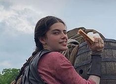 a woman is holding something in one hand and looking at the other while standing next to an old wooden barrel