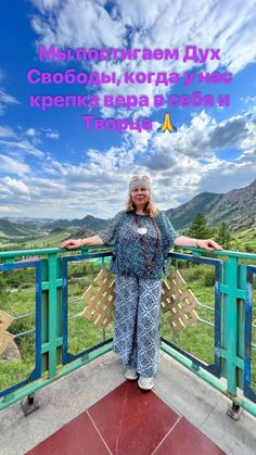 a woman standing on top of a balcony next to a lush green field with mountains in the background