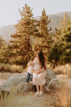 a woman holding a baby sitting on top of a rock in the middle of a forest