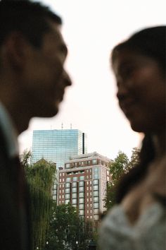 two people standing next to each other in front of tall buildings with trees and water