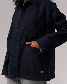 The Recycled Cotton Denim Jacket is a heavy-weight denim chore coat. The jacket features a folded collar, button front, pocket with a pen holder on the left breast, buttoned size adjusters on the back, and dual wraparound pockets at the waist on each side. The material is a 12.6oz recycled denim with reinforced stitching. Chore Coat, A Pen, Recycled Denim, Black Denim Jacket, Pen Holder, Recycled Cotton, Heavy Weight, Front Pocket, Denim Jacket