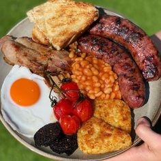 a person holding a plate with sausage, eggs, beans, toast and tomatoes on it