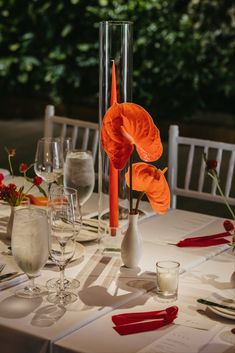 an orange flower is in a tall vase on a table set with wine glasses and place settings