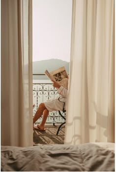 a person sitting on a chair reading a book in front of a window with white curtains