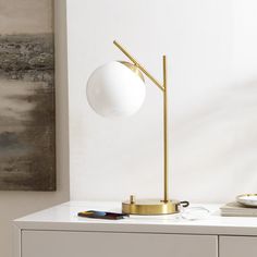 a white table with a gold lamp on it and a painting hanging in the background