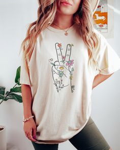 Looking for a shirt that will show your support for peace and love? Look no further than the peace sign shirt! This shirt is perfect for anyone who wants to express their love for peace and happiness. The shirt features a hand giving the peace sign surrounded by wildflowers making it a great choice for any fashion-savvy individual. Please not that this T-shirt is a Standard Unisex fit, if you would like an oversized look then size up 1 to 3 sizes. Comes in multiple colors! ✨️SIZE AND FIT: Your s Cotton Peace Sign Short Sleeve T-shirt, Hippie T-shirt With Screen Print For Spring, Summer Crew Neck Top With Peace Sign, Spring Hippie T-shirt With Screen Print, Cotton Short Sleeve T-shirt With Peace Sign, Hippie Style Spring T-shirt With Screen Print, White Hippie Crew Neck T-shirt, White Crew Neck Hippie T-shirt, Casual Cotton T-shirt With Peace Sign