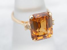 This mounting dates to the Retro era, although we've replaced the center gemstone. The design is lovely, and simple but graceful. We've set this vintage mounting with a sparkling, beautiful golden zircon! This stone is from Sri Lanka, the source of some of the most richly colored, brilliant zircons in the world. Natural zircon was a common sight in the jewelry stores of the 1930s, in fact, it's one of the original birthstones for December, but nowadays it's been overshadowed by cubic zirconia. Similar name, but the two stones are not related in any way.Metal: 14K Yellow GoldGem: Orange ZirconGem Measurements: 12.6 x 10.1 mm, Emerald CutAccents: 2 Diamonds totaling .09 Carats, VS in Clarity, I in ColorRing Size: 6.50Marks: “14K” Stamped on the inside band Cut Orange, Retro Era, Deco Blue, Solitaire Pendant, Blue Zircon, December Birthstone, Color Ring, The Source, Eternity Bands