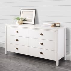 a white dresser with two pictures on top and a potted plant in the corner