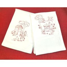 two white towels with drawings on them sitting on a red tablecloth, one is folded and the other has an image of a house