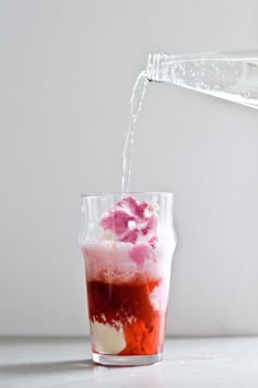 a red drink being poured into a glass filled with ice and water on a white surface