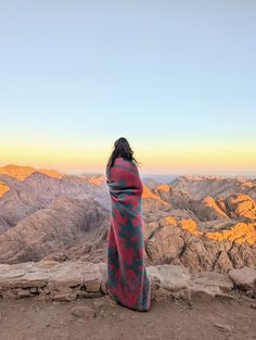 a woman wrapped in a blanket sitting on top of a mountain looking at the mountains