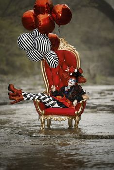 a clown sitting on a red chair in the water with balloons attached to it's back